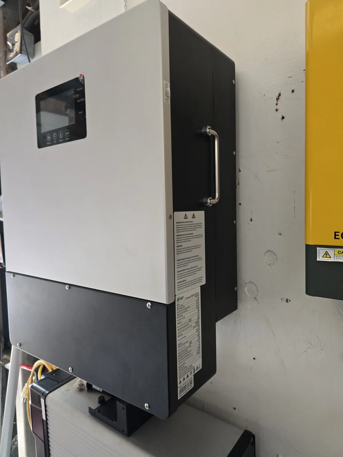 Angled view of a white-and-black wall-mounted hybrid inverter with a front display, side handle, and warning labels.