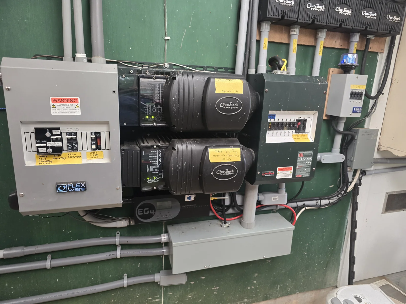 Wall-mounted dual OutBack inverter installation with adjacent electrical enclosures, charge controllers, conduit, and labeled wiring.